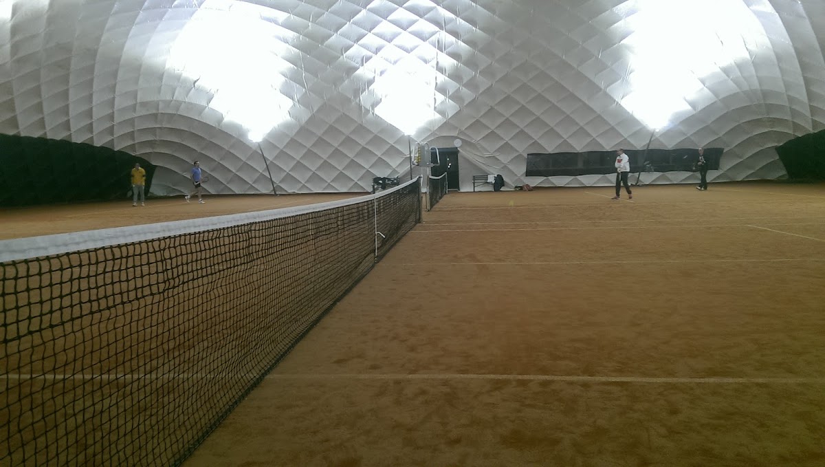 Overdekte padelbaan met bruinrood zand, net op voorgrond, LED-verlichting en witte koepeldak.