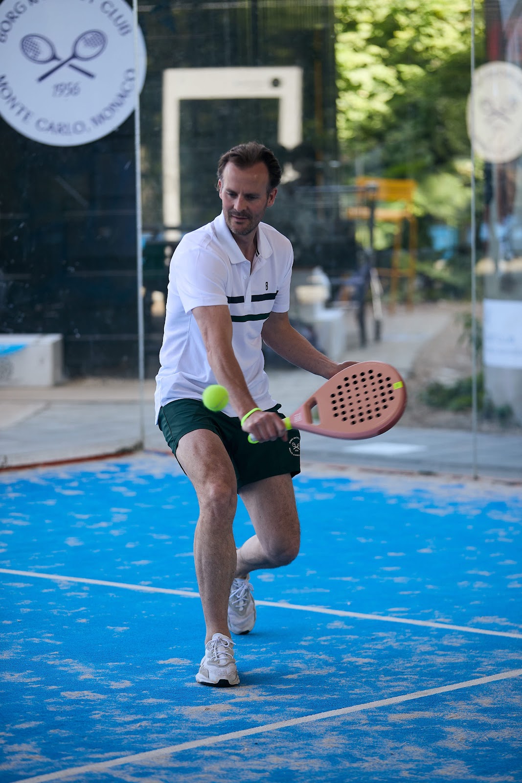 Padelspeler in witte polo op blauwe padelbaan met glazen wanden en groene bal