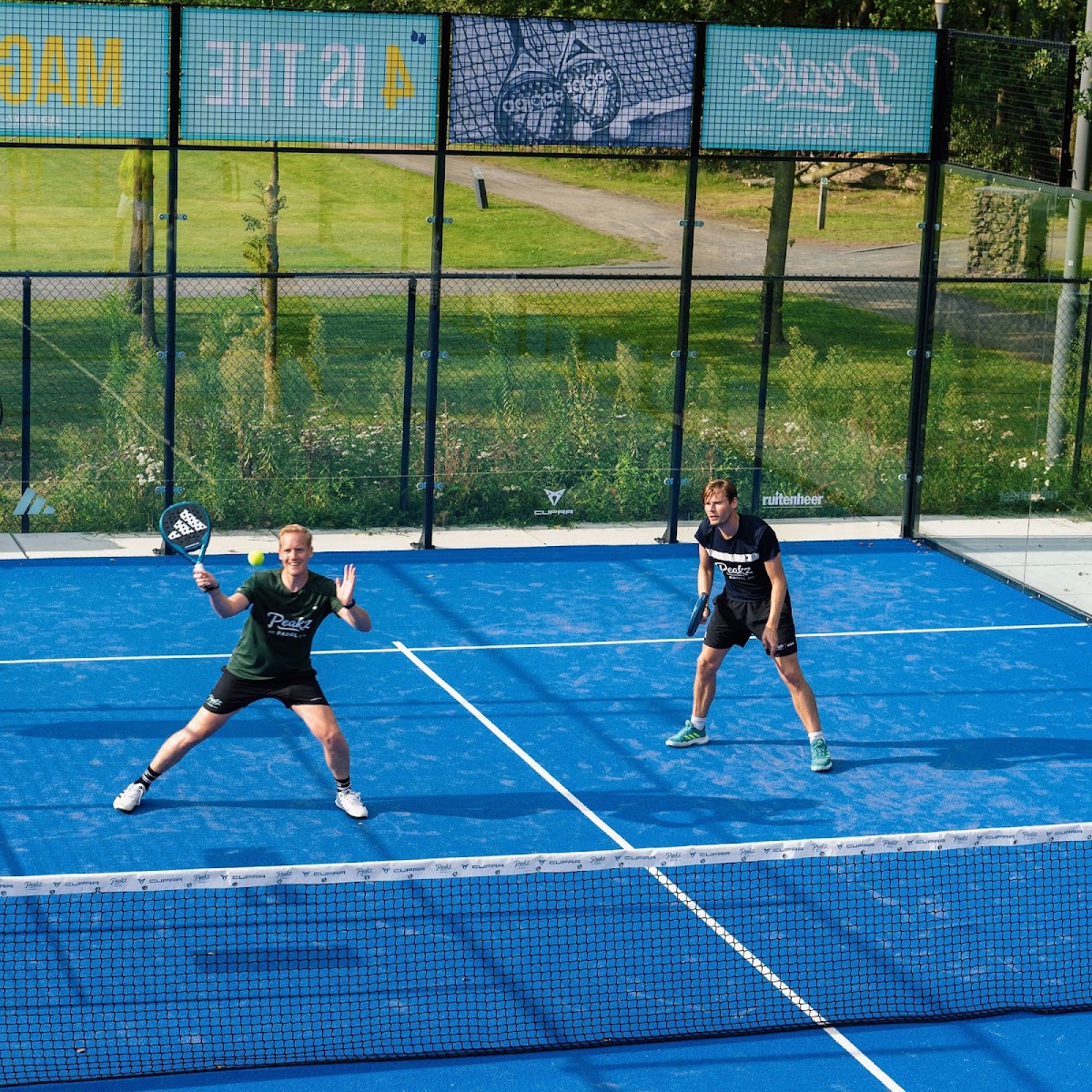 Twee spelers op een blauwe padelcourt met mesh-omheining en groene achtergrond.
