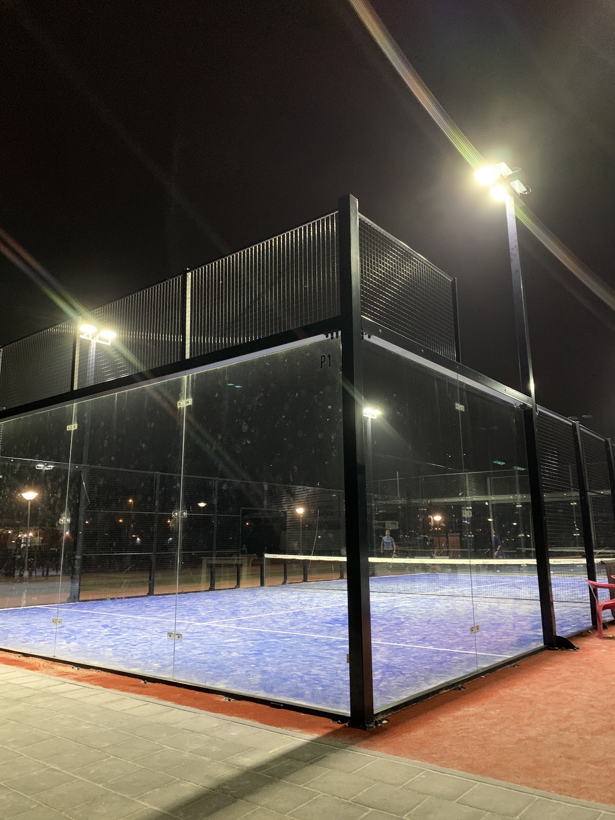 Verlichte outdoor padelbaanmet blauwe QuickPadel-ondergrond, volledig omsloten door glas en metalen mesh.