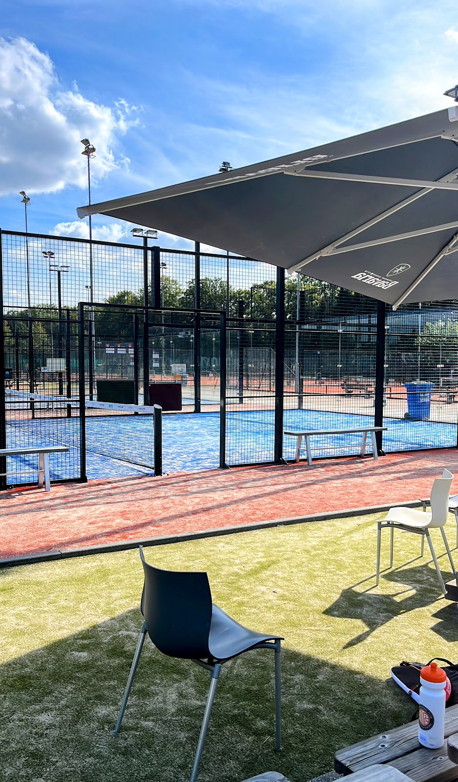 Outdoor padelclub met blauwe QuickPadel-banen, zwarte omheining en parasol onder blauwe hemel