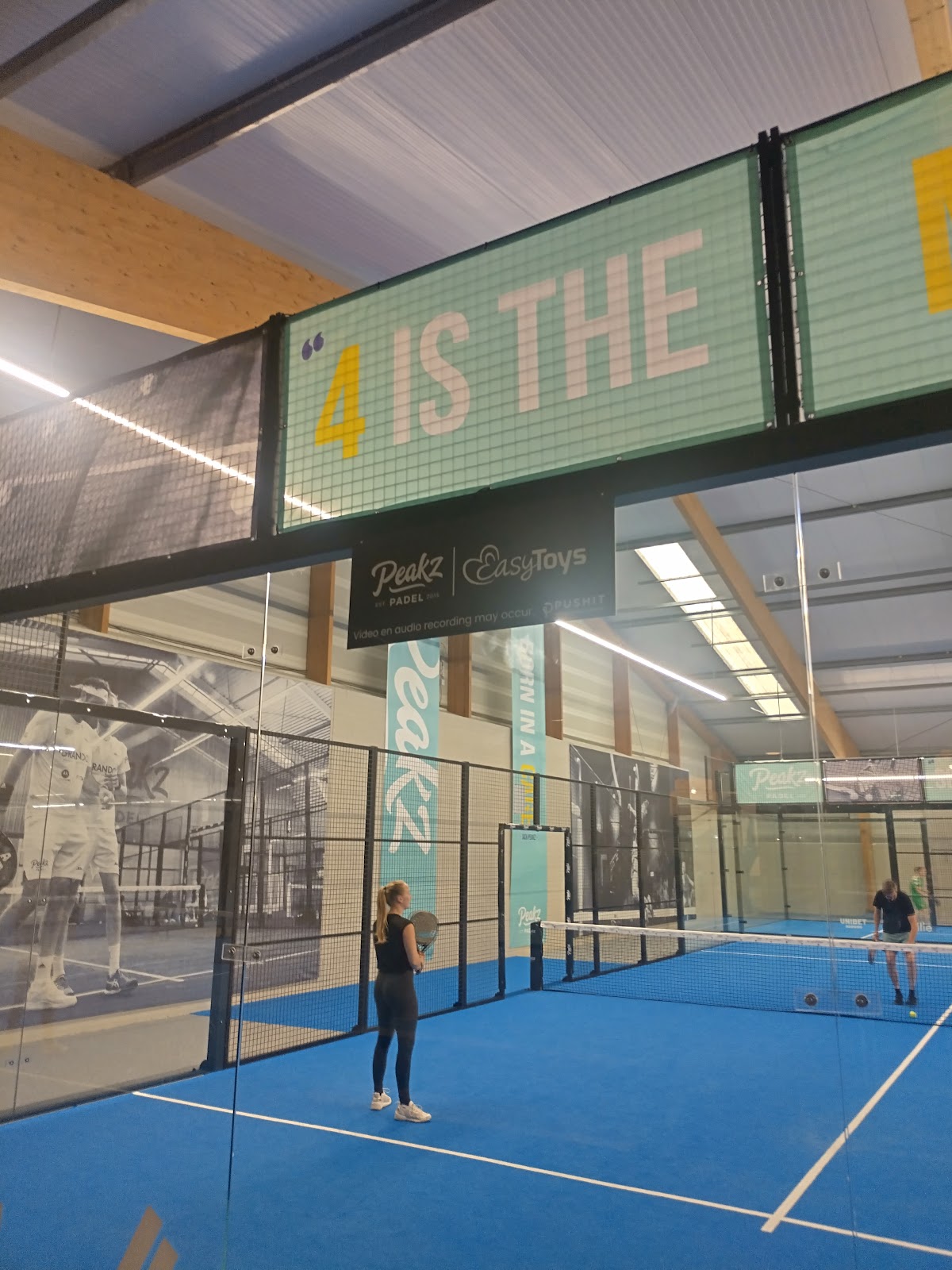 Indoor padelclub met blauwe hardcourt, spelers in actie, groene signage '4 IS THE'