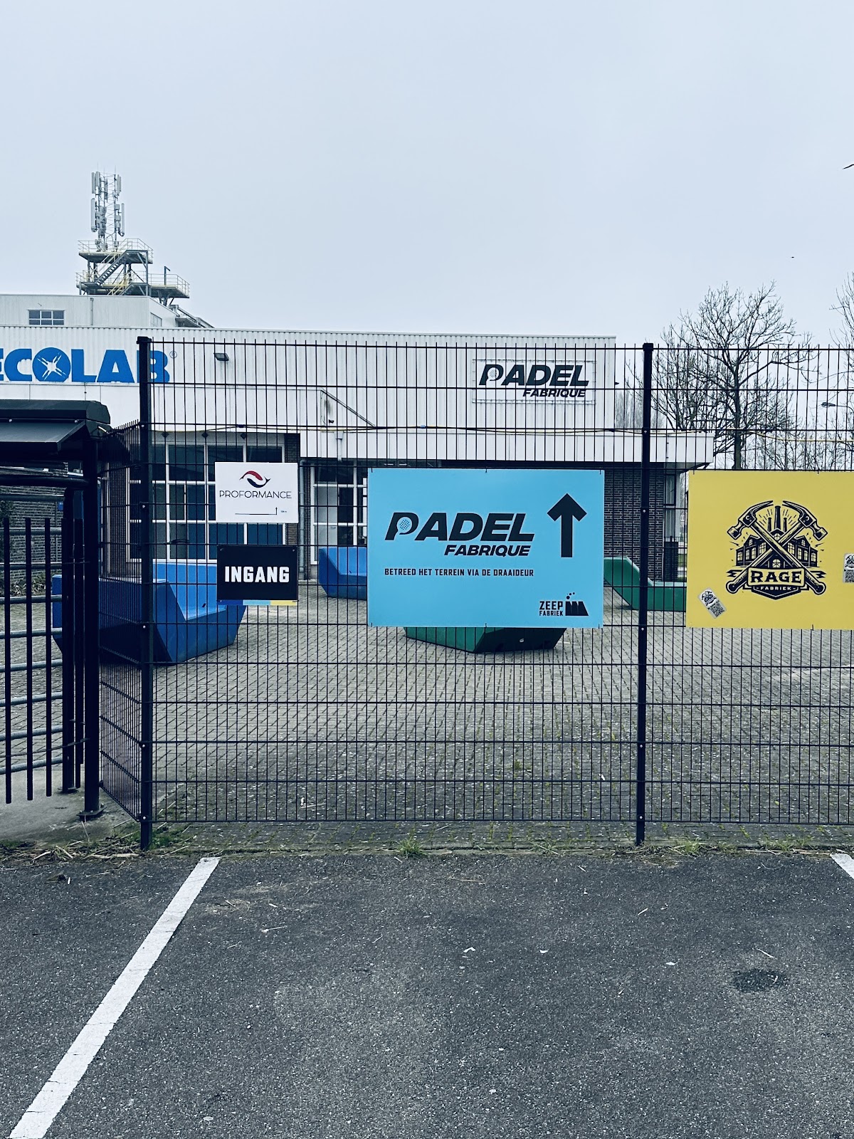 Ingang van Padel Fabrique met blauw en geel bord, omheind terrein