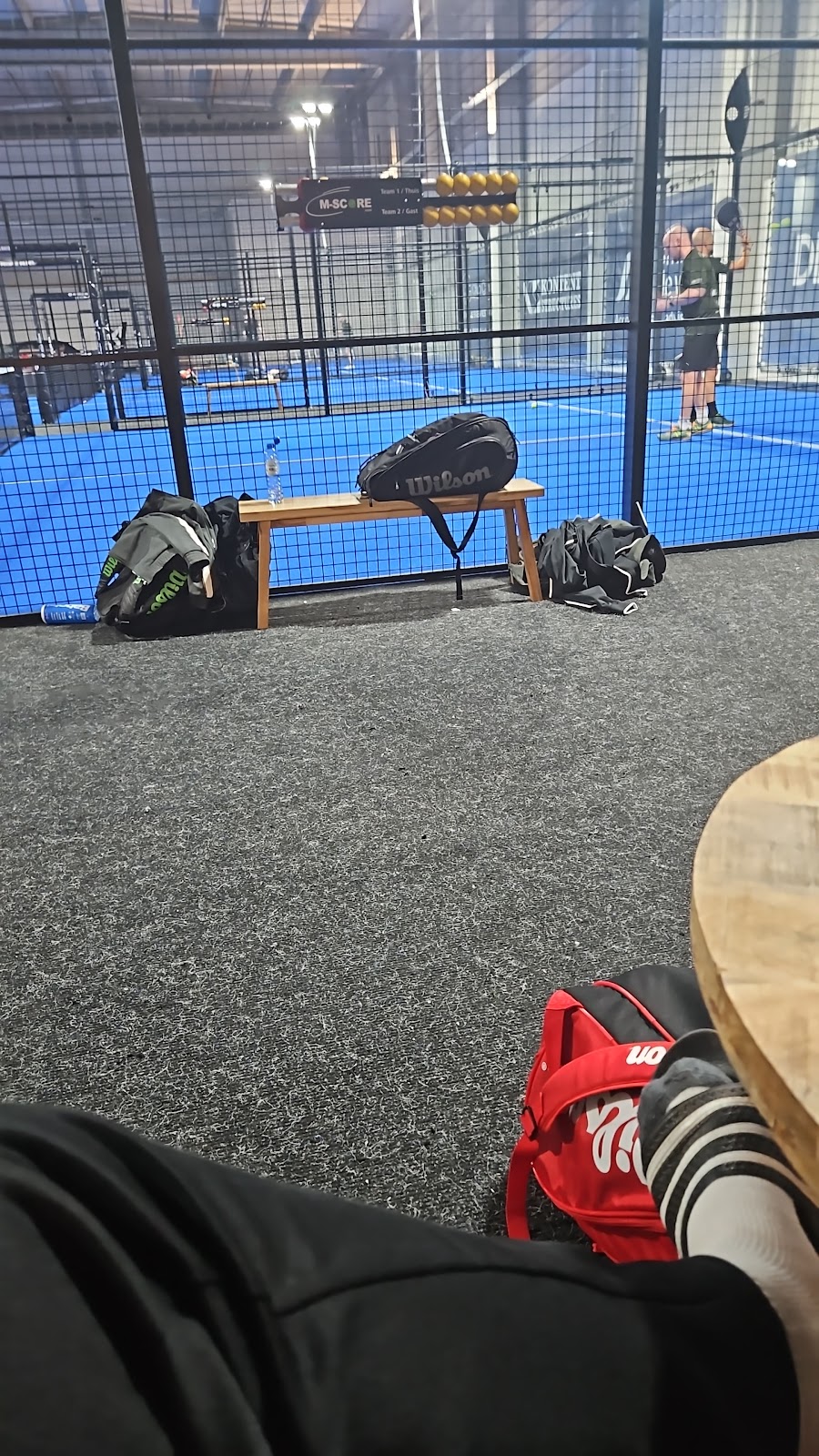 Binnenpadelclub met blauwe velden, tassen op houten bank en floodlights