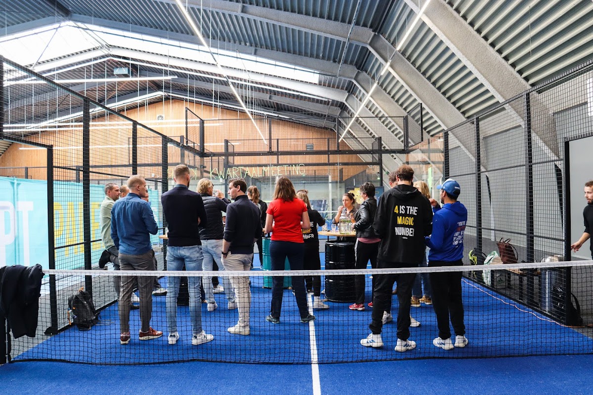 Padelbaan met blauwe ondergrond en groep mensen bij hospitality area in overdekte hal