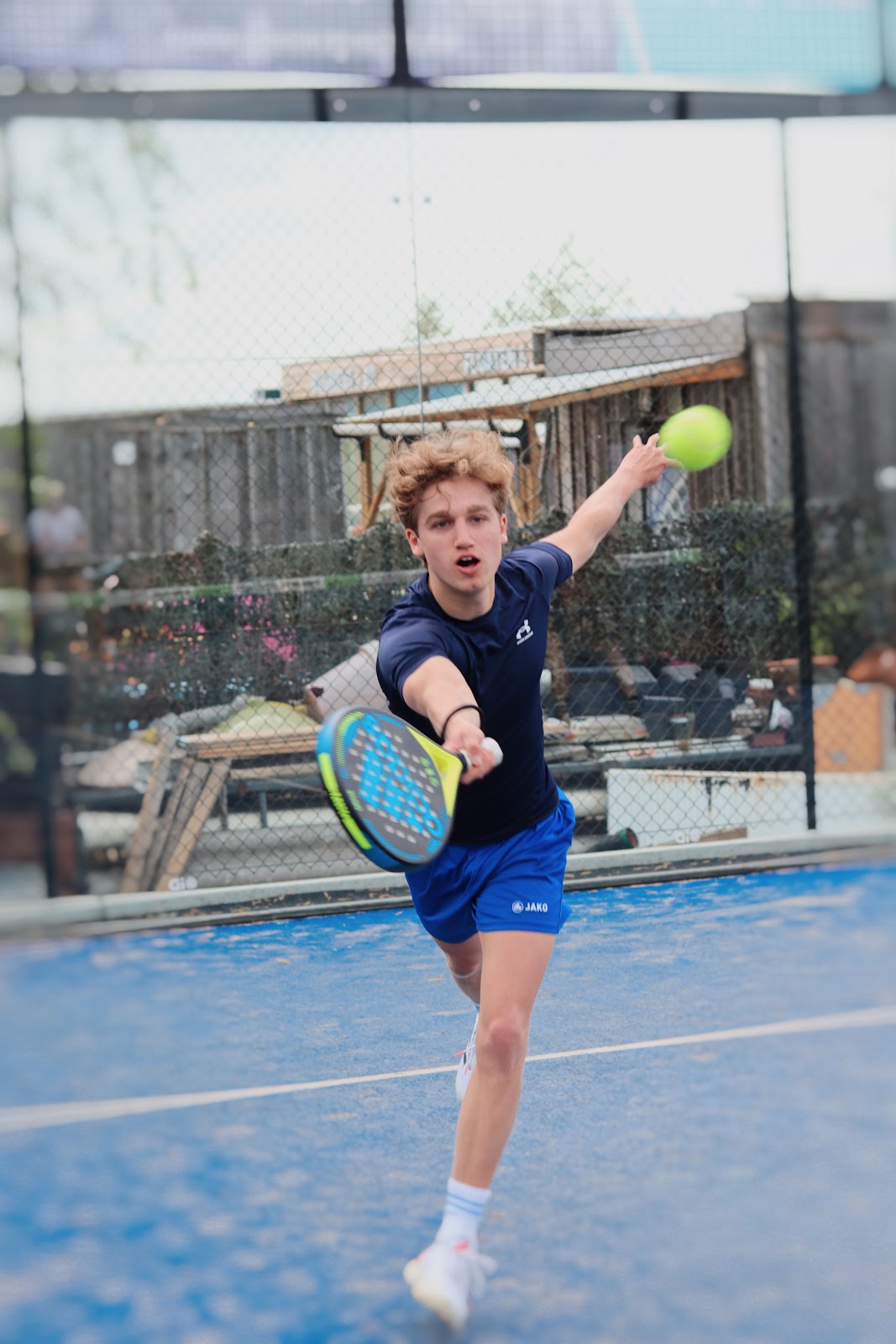 Jongespeler serveert op blauwe padelbaan met geel balletje tijdens outdoor match