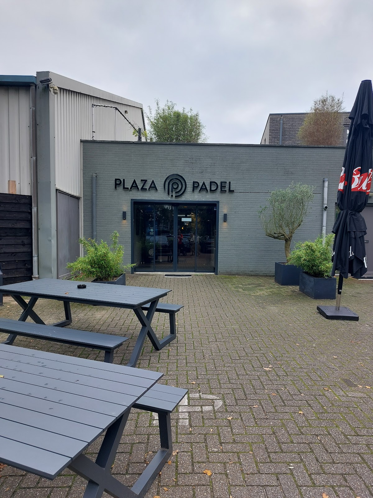 Plaza Padel clubgebouw met terras, picknicktafels en ingangen zichtbaar