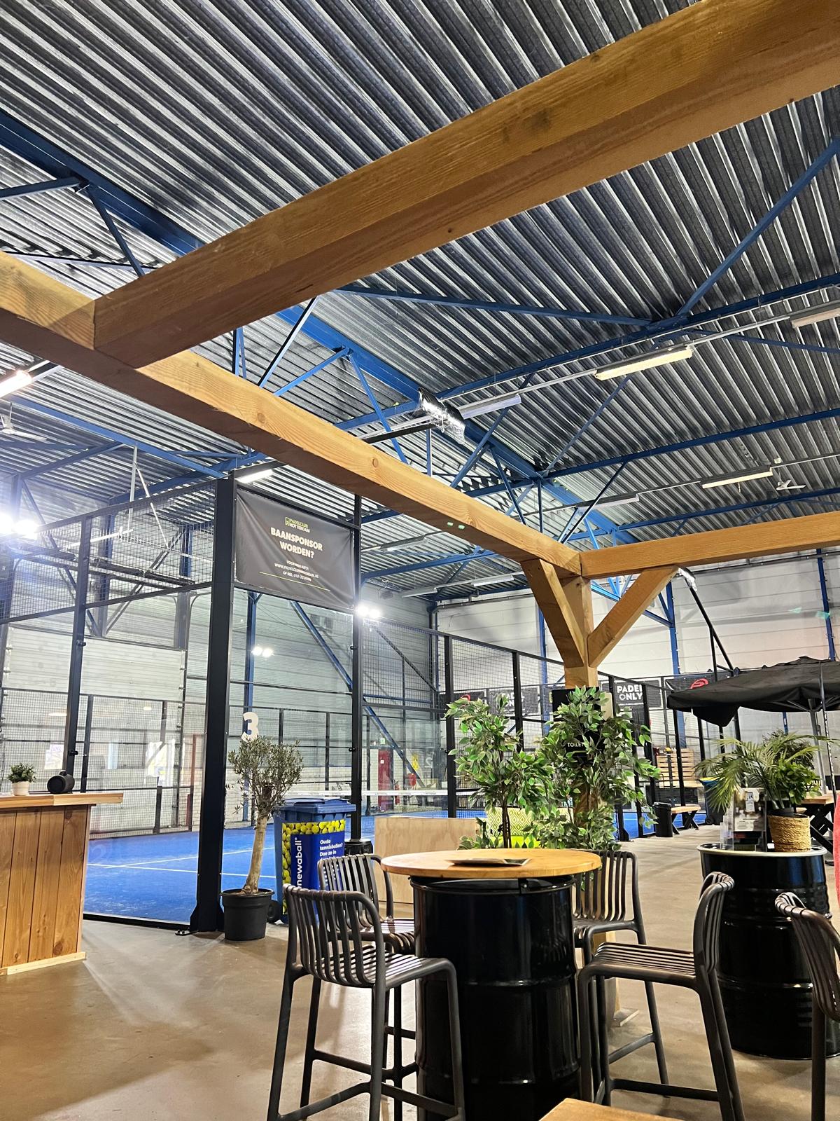 Padel club met blauwe court, houten balken, bar en zithoek onder golfplaten dak