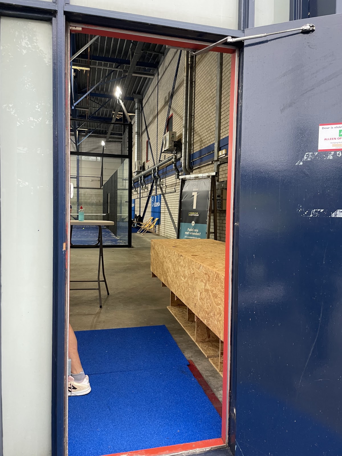 Ingang padelbaan met blauwe mat, houten balie en overdekt sportcomplex