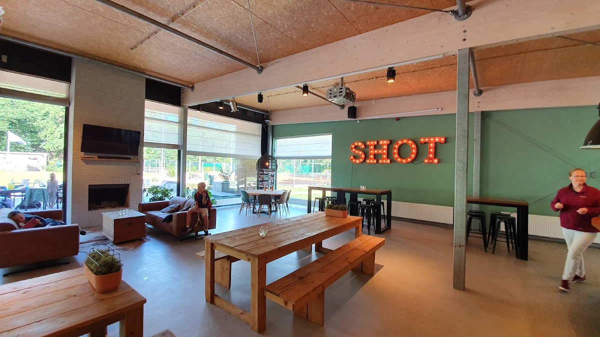 Moderne padelbaan met houten tafels, groene wand met SHOT-letters en zitgedeelte