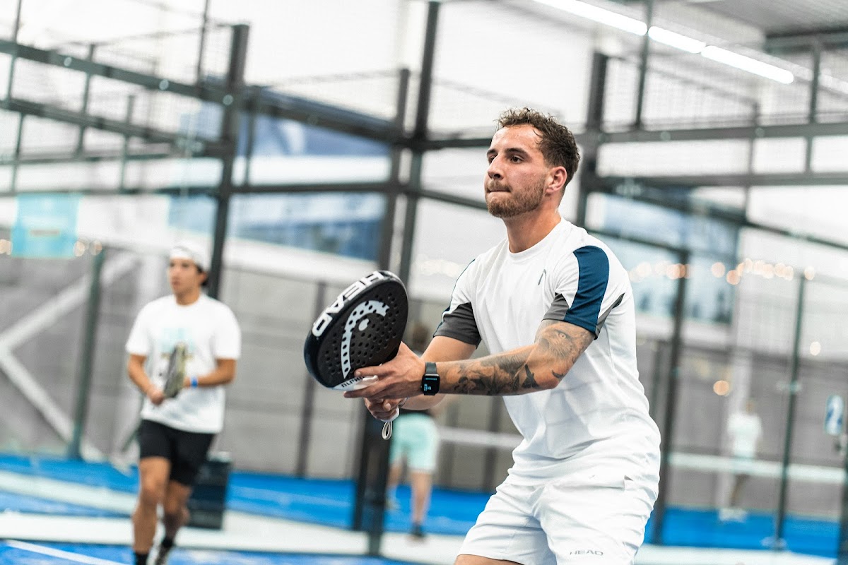Padelspeler in actie op blauw kunstgras court met glazen omheining en daglicht verlichting