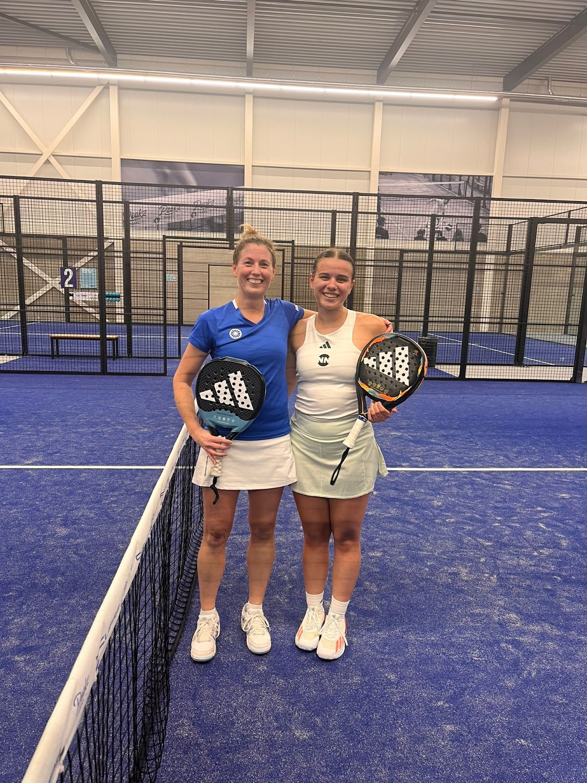 Twee speelsters poseren met padel rackets op een binnenbaan met blauwe ondergrond en zwart gaas omheining.