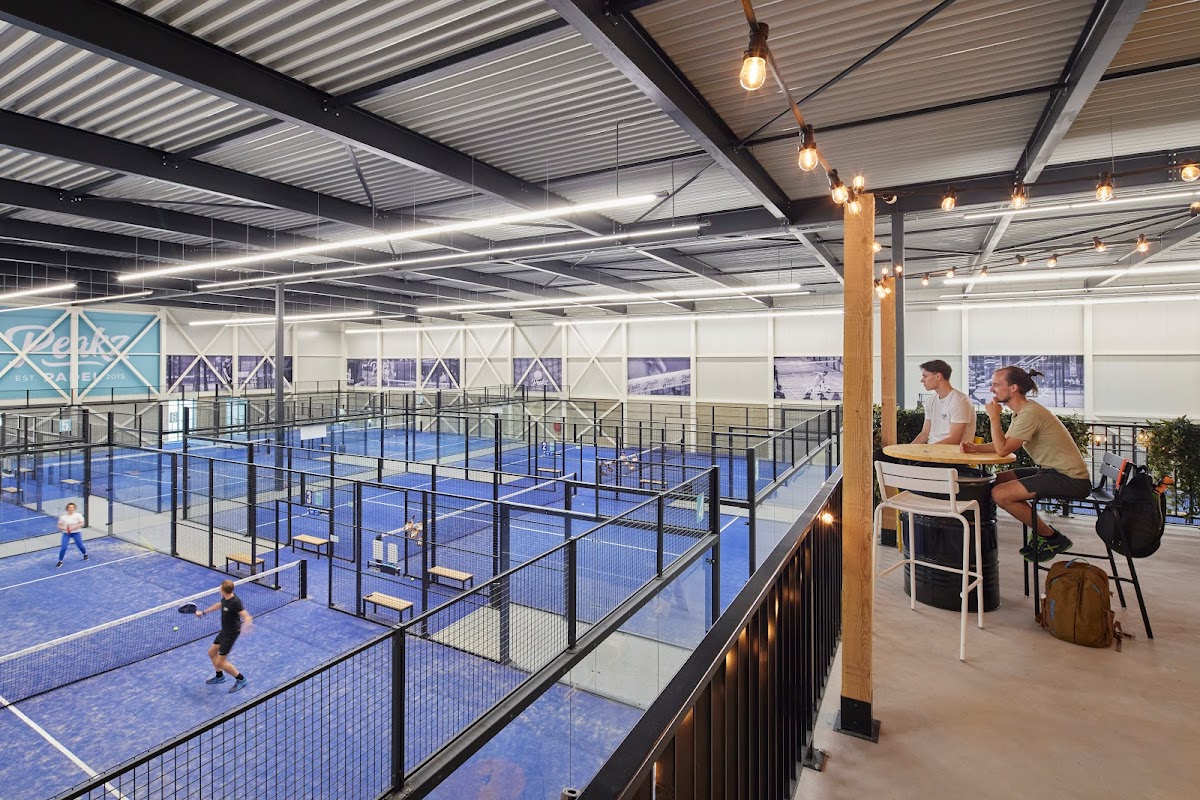Moderne indoor padelclub met meerdere courts, blauwe velden, glazen wanden, verlichting en spectatorentribune.