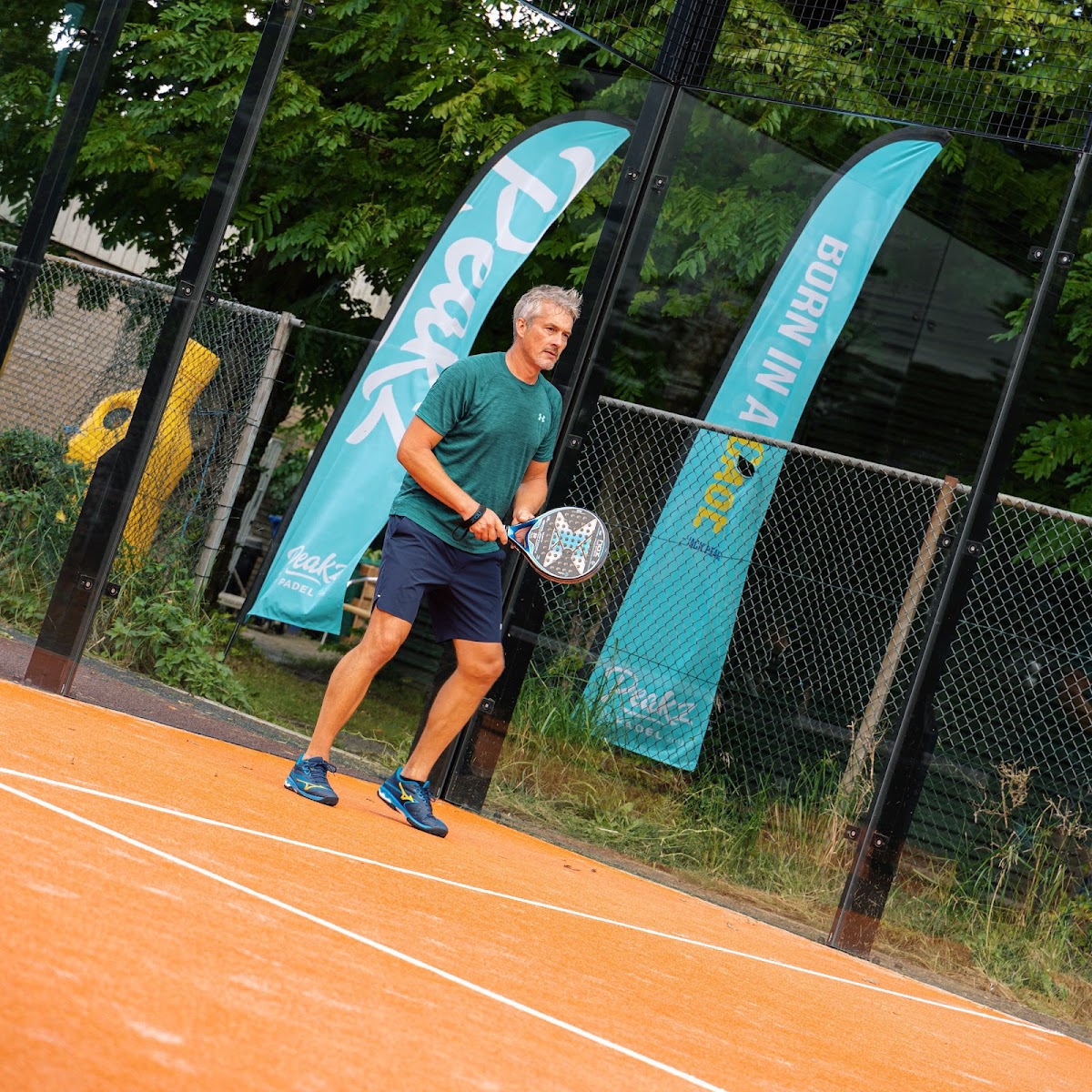 Padel speler op oranje baan met turquoise banners en groene omheining.