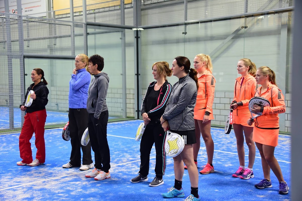 Groep vrouwen op blauwe padelbaan in overdekte sporthal met glazen wanden