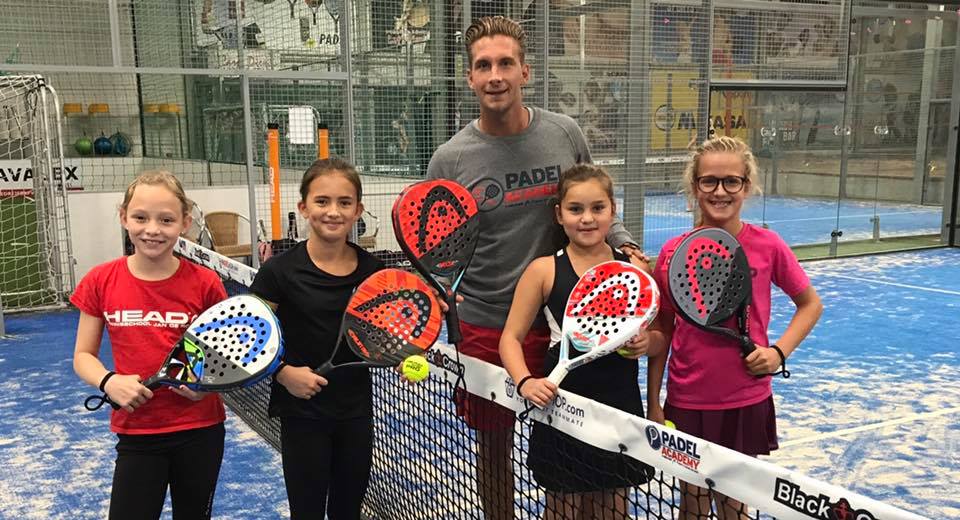 Padeltrainer met vijf kinderen op een indoorbaan met blauwe ondergrond, allemaal met padelrackets.