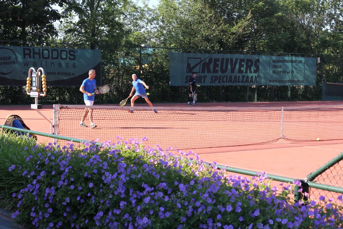 Padelbaan met spelers in blauwe shirts, paarse bloemen op de voorgrond en bomen op achtergrond