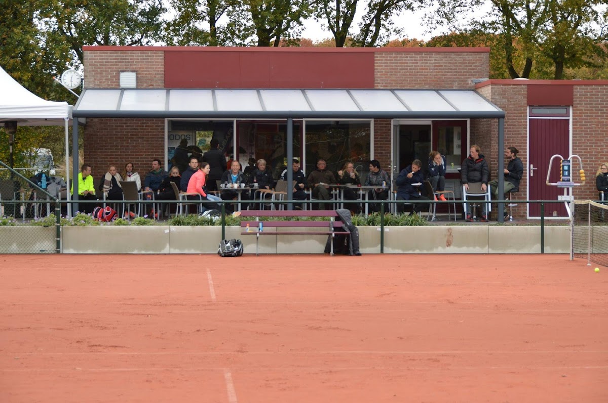 Padelbaan buiten met clubhuis, toeschouwers op terras, rode ondergrond zichtbaar.
