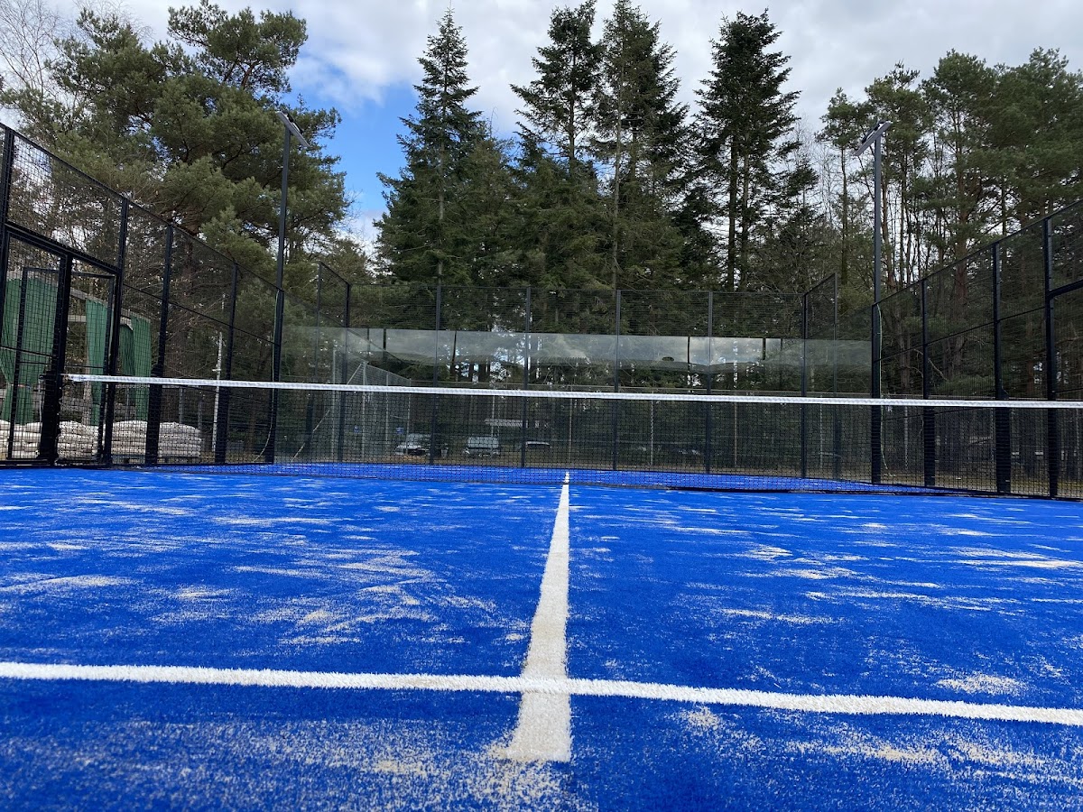 Buitenpadelbaanmet blauwe QuickPadel-oppervlak, witte lijnen, omsloten door metalen gaas en glaswand.