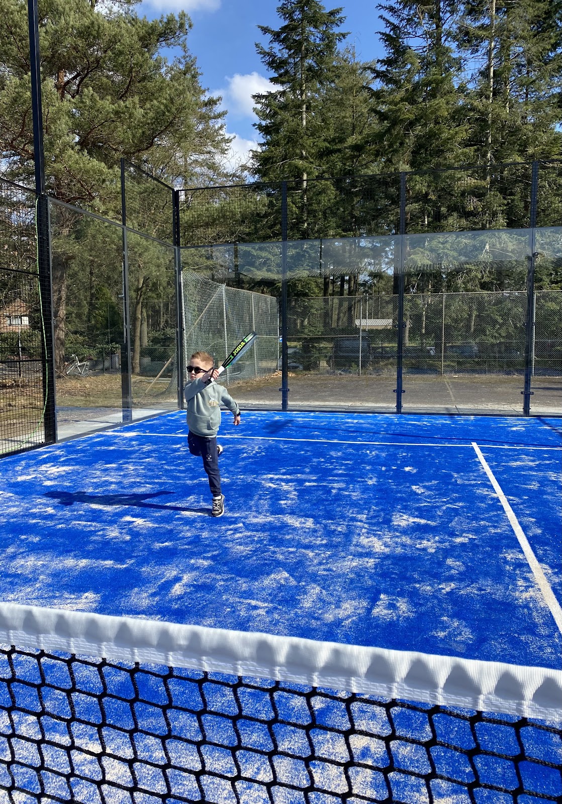 Kind speelt padel op blauw hardcourt met glazen wanden in buitenbaan