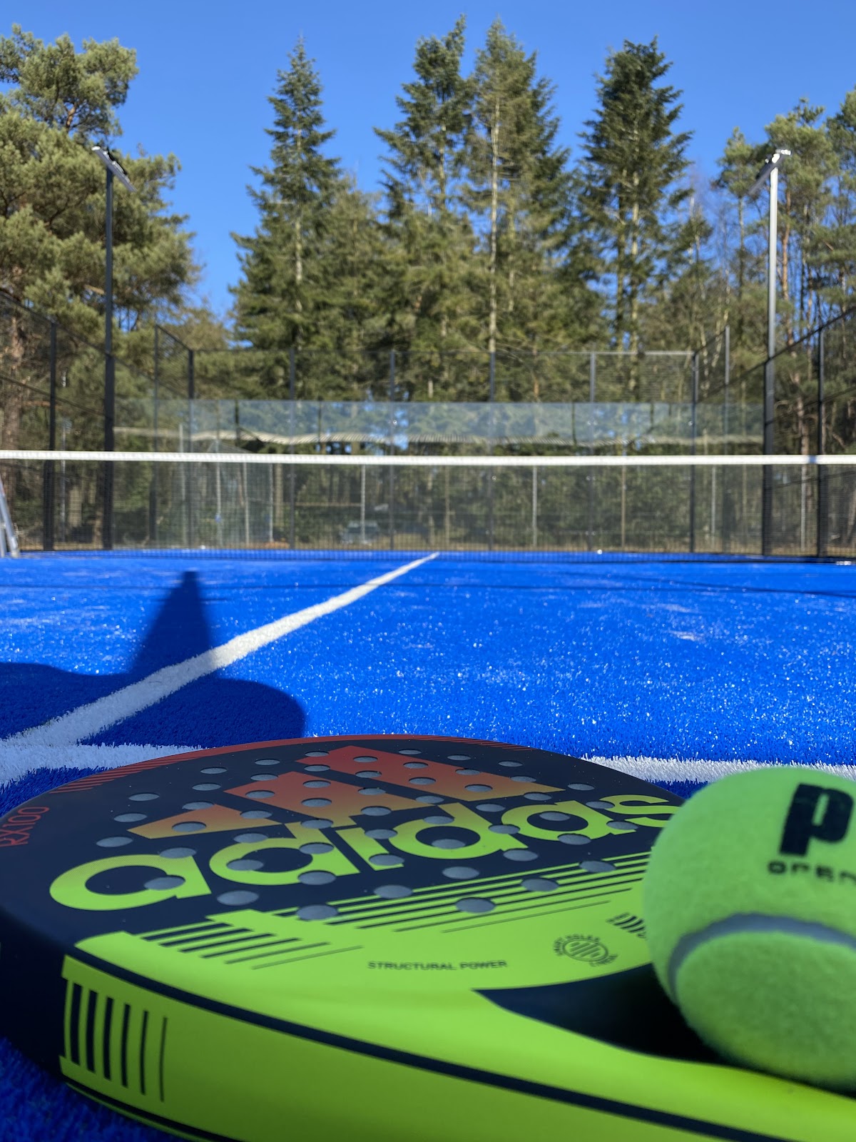 Padelbaan met blauwe kunstgrasoppervlak, omgeven door gaas en glazen wanden, groene paddle in voorgrond.