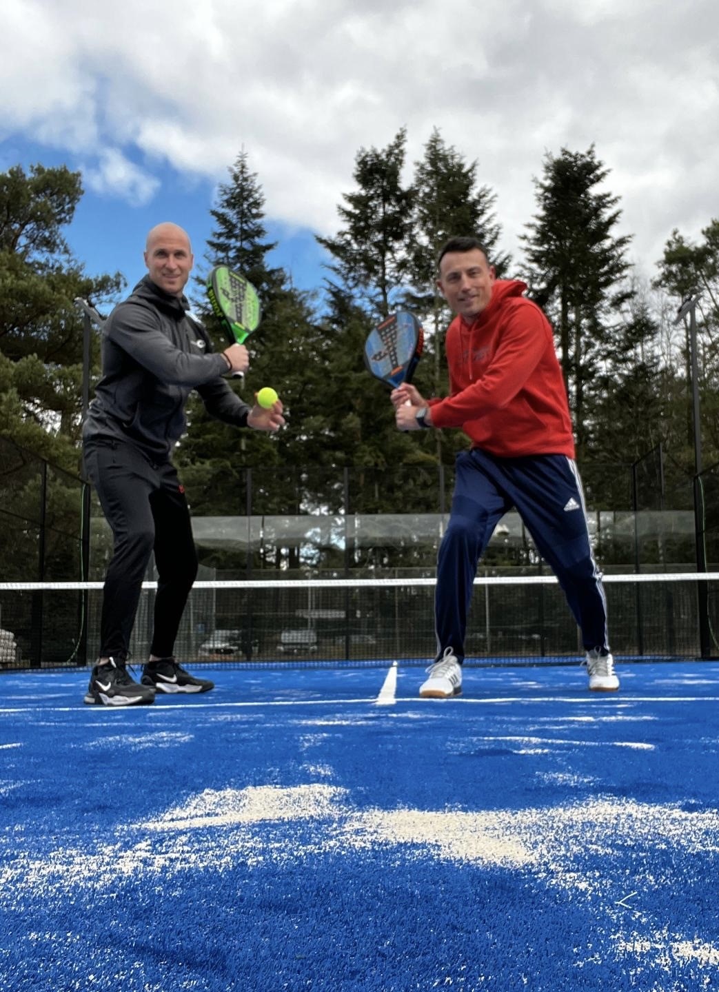 Twee spelers op een blauwe padelcourt met glazen achterwand en bosachtergrond.