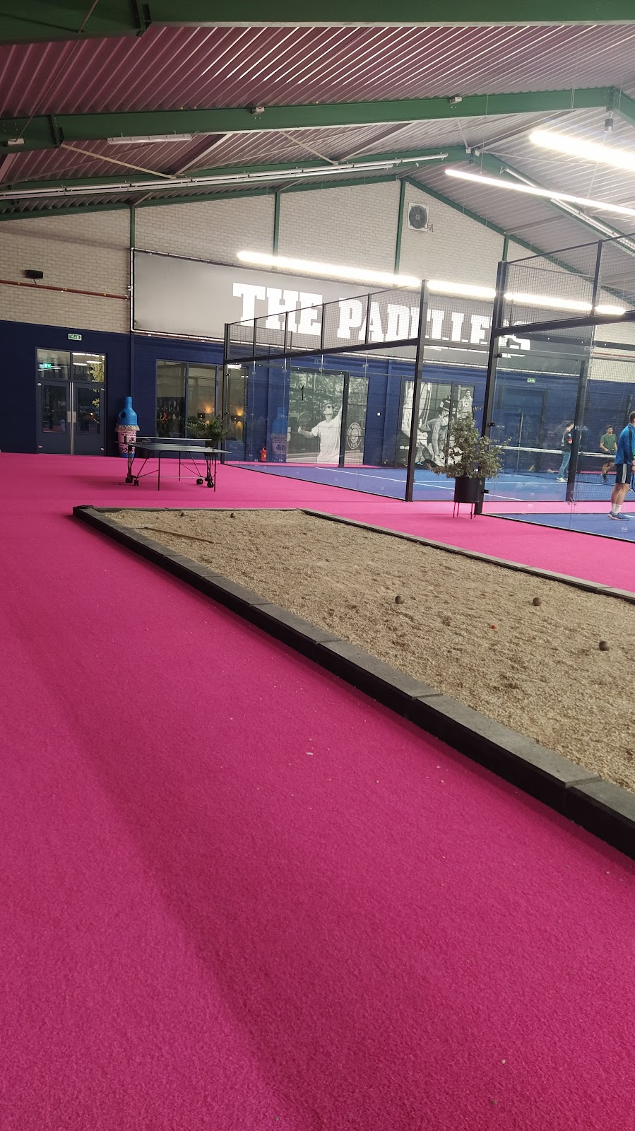 Binnenpadelclub met roze velden, zandvak en blauwe ramen onder industrieel dak