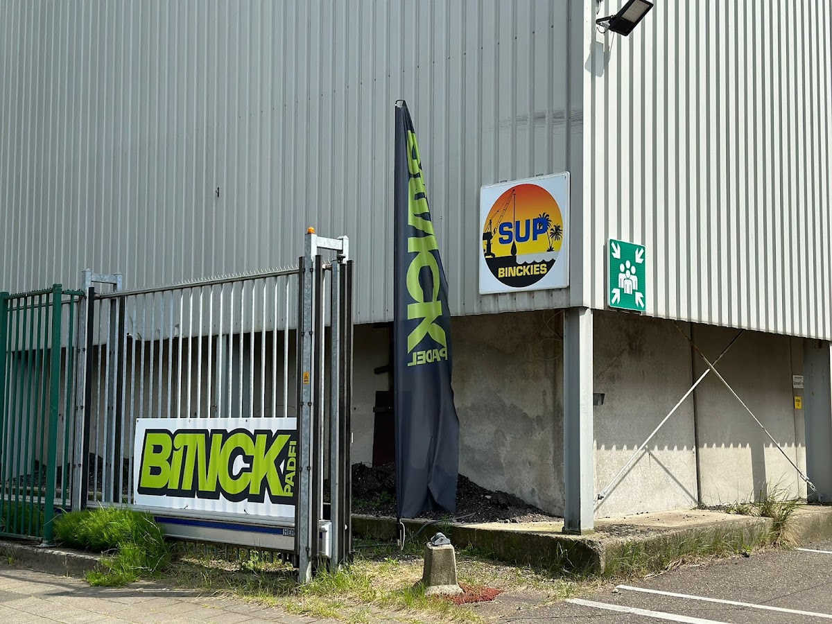 Ingang van Binckies padel club met groen hek, bord en opgeslagen surfboard