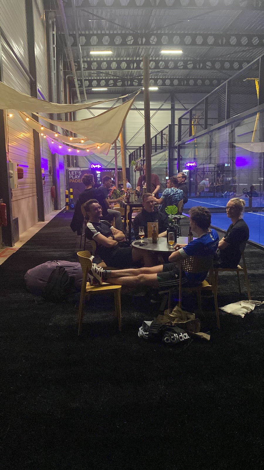 Avondsfeer op padelbaan met loungezitjes, paarse verlichting en speelveld op achtergrond