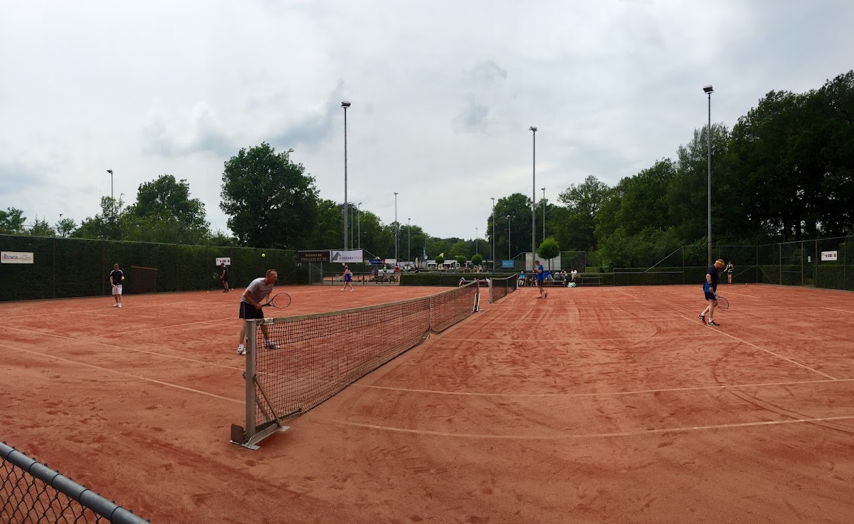Outdoor padelclub met meerdere rode gravel banen, verlicht door hoogtelampen, omgeven door groen.