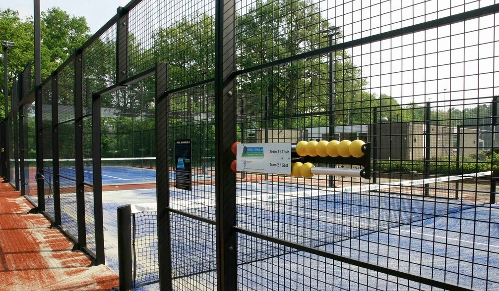 Outdoor padelbaancomplex met blauwe hardcourts, zwarte omheiningen en gele trainingsballen.
