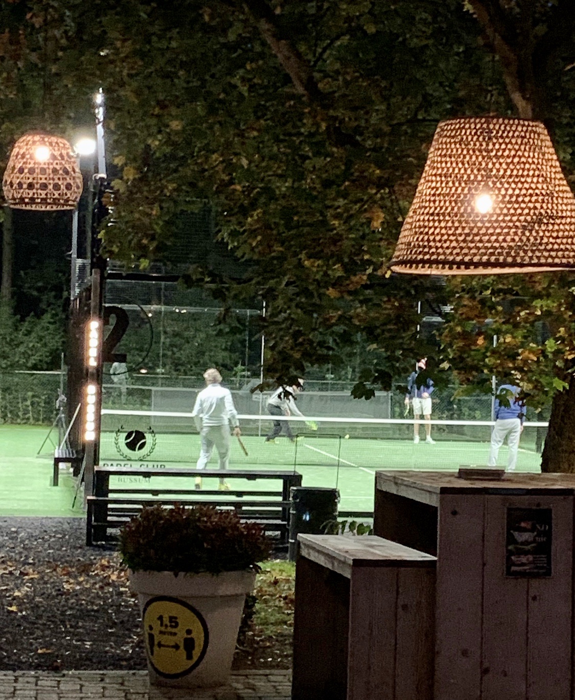 Avondelijk padelbaan met kunstgras, verlicht door grote hanglampen en floodlights, met spelers in actie.