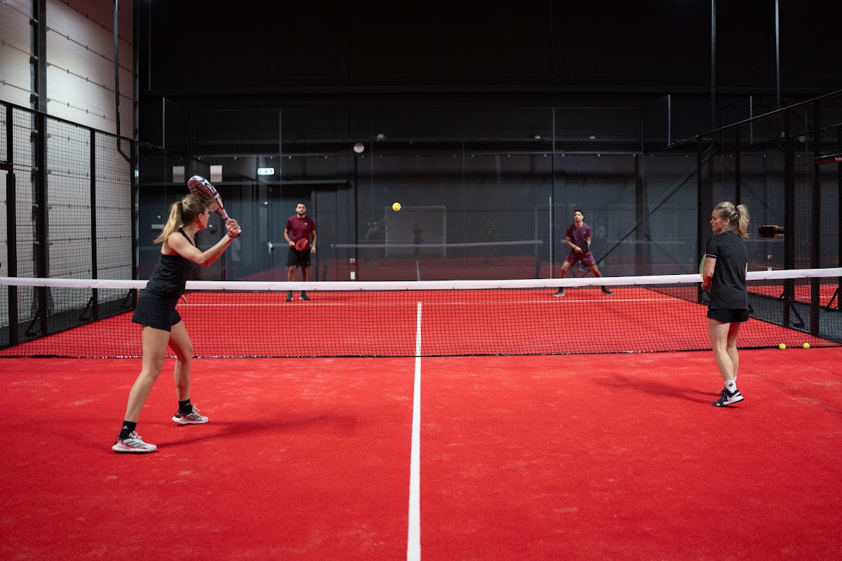 Twee speelsters spelen padel op een rood kunststofveld met netwerk omheining in overdekte baan.