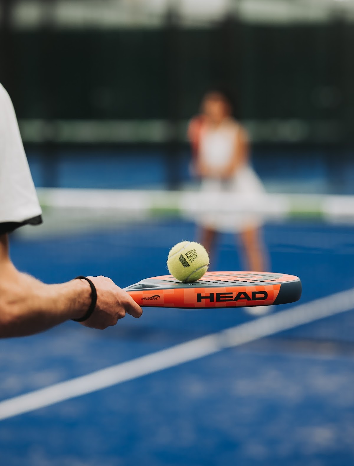 Padelracket met bal op blauw hardcourt, speler op achtergrond