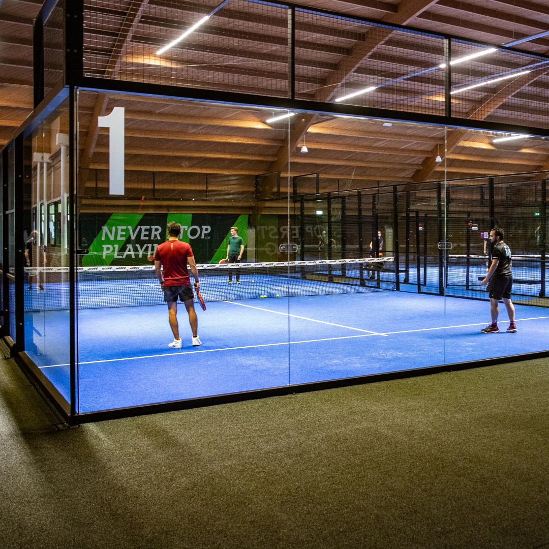 Moderne indoor padelbaan met blauwe vloer, zwarte wanden en LED-verlichting. Spelers in actie op professionele court.